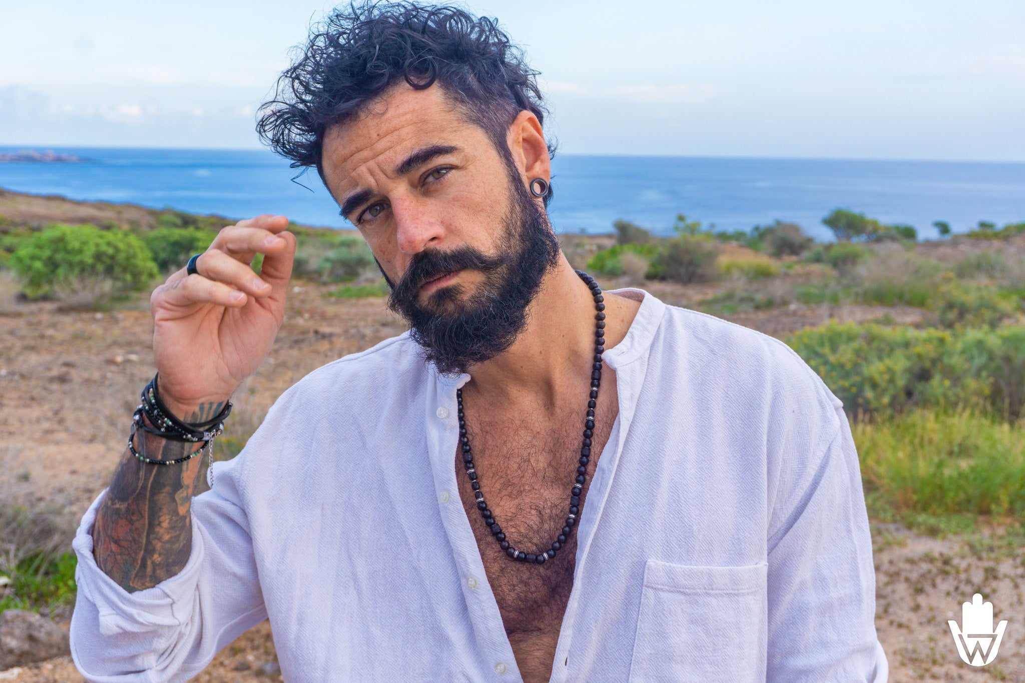 A man with a beard and curly hair wearing a white button-down shirt and a beaded necklace, standing outdoors with a coastal landscape in the background.