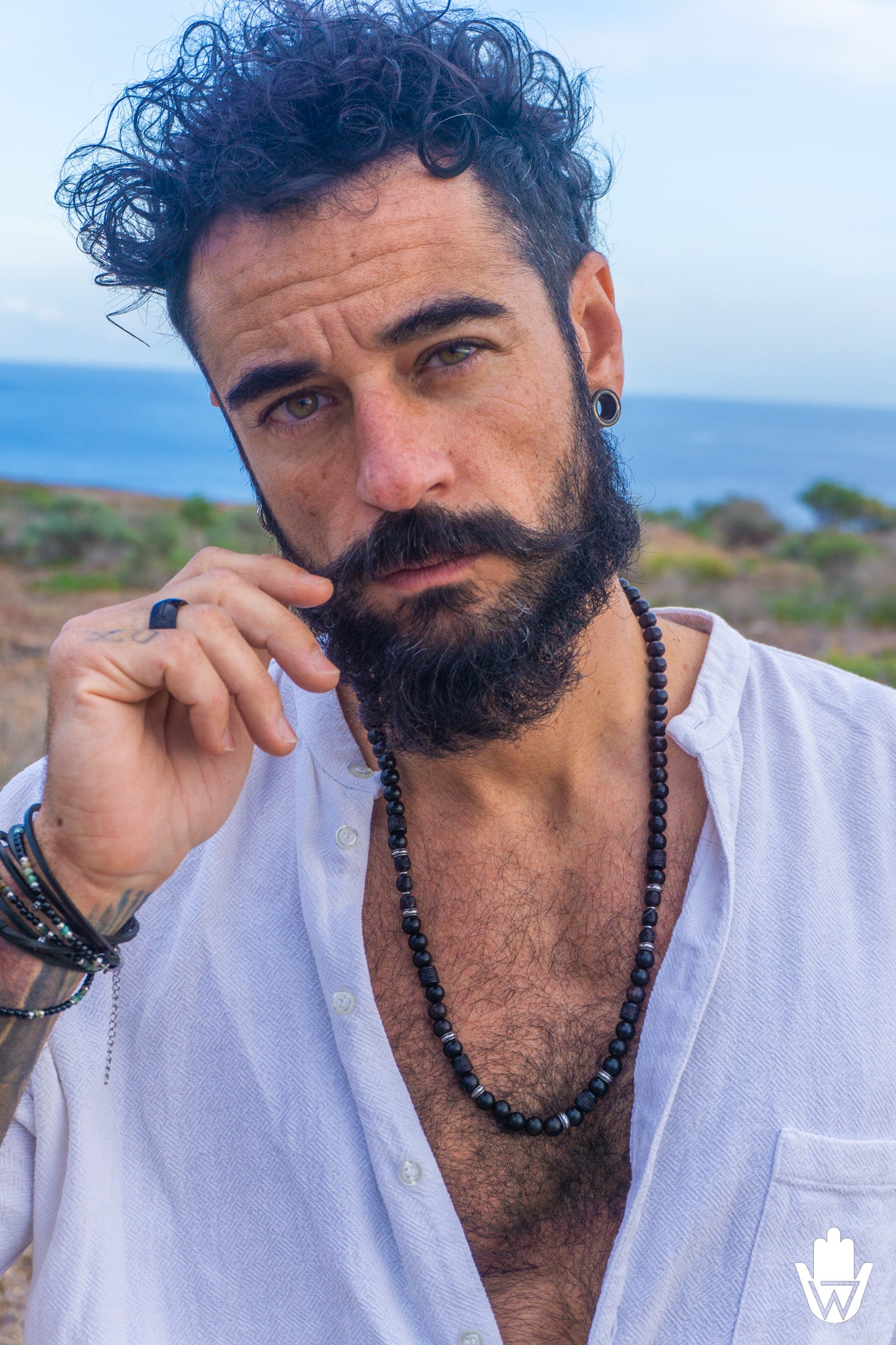 A close-up portrait of a man outdoors with the ocean in the background, wearing a white open shirt and a long black beaded necklace.