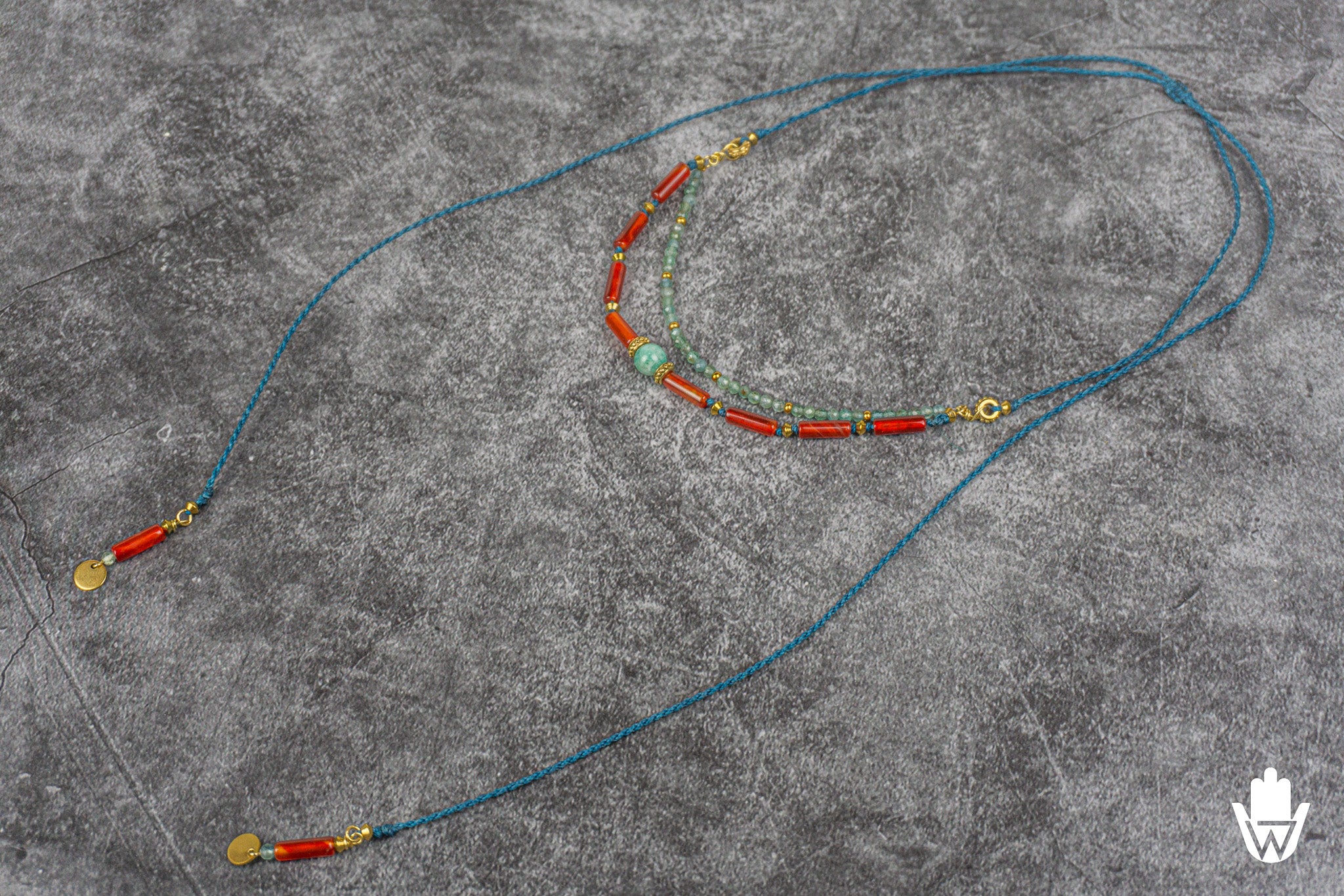 A bohemian-style beaded necklace with a turquoise center bead, red cylindrical beads, and gold-toned metal accents, laid out on a textured gray surface.