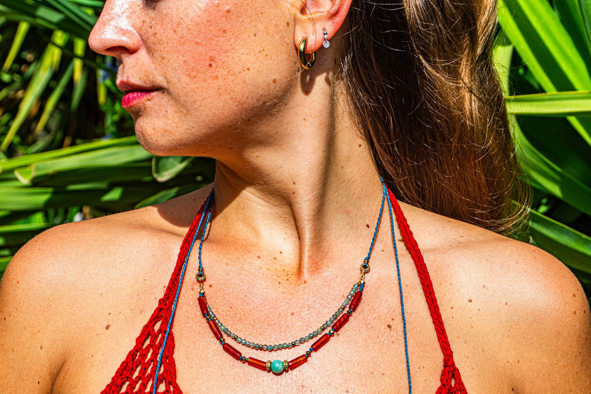 A close-up lifestyle shot of a woman wearing a bohemian-style beaded necklace with red, blue, and turquoise beads, set against a background of green tropical leaves.