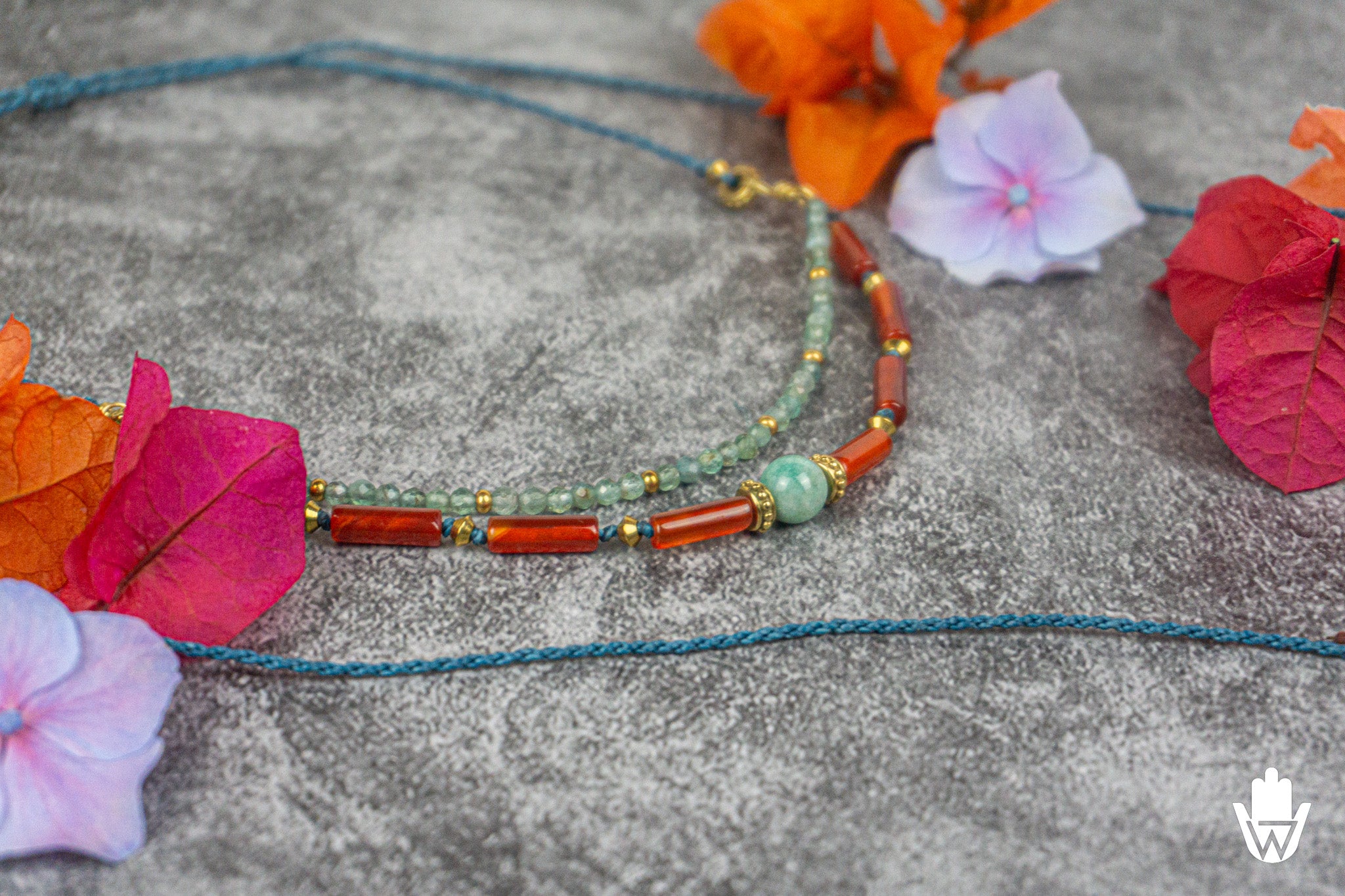 A beaded necklace with turquoise-blue cord, orange-red cylindrical beads, pale green round beads, and small gold spacer accents, arranged on a textured gray surface with colorful flowers around it.