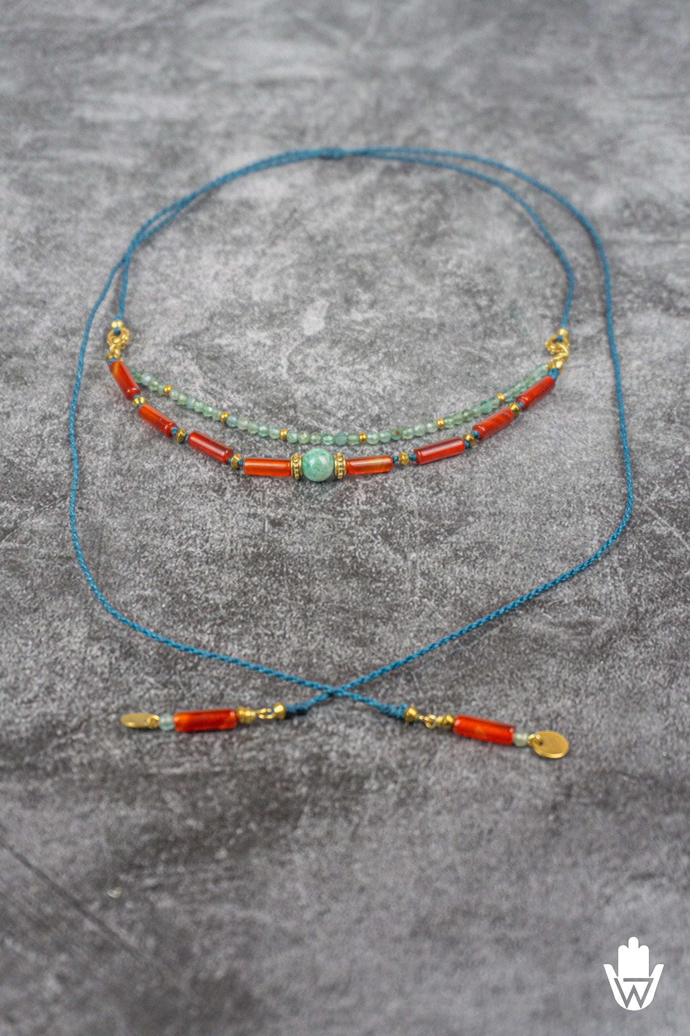 A double-strand beaded necklace with turquoise blue cords, orange-red cylindrical beads, light green round beads, and gold-toned spacer details, displayed on a textured gray surface.
