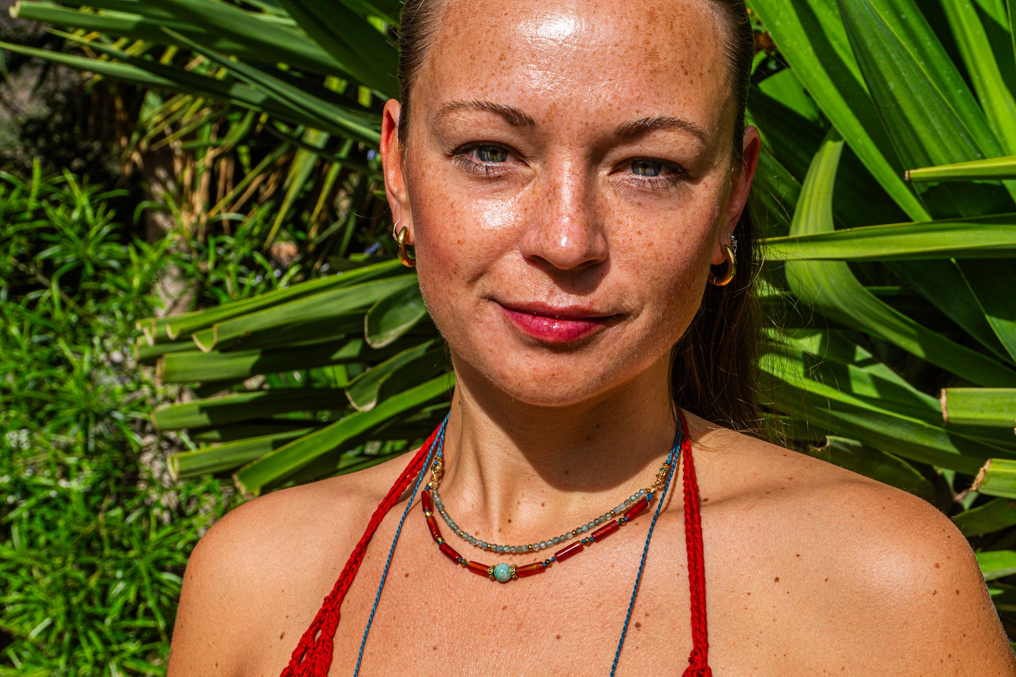 A close-up portrait of a woman outdoors in front of green tropical leaves, wearing a layered choker necklace i