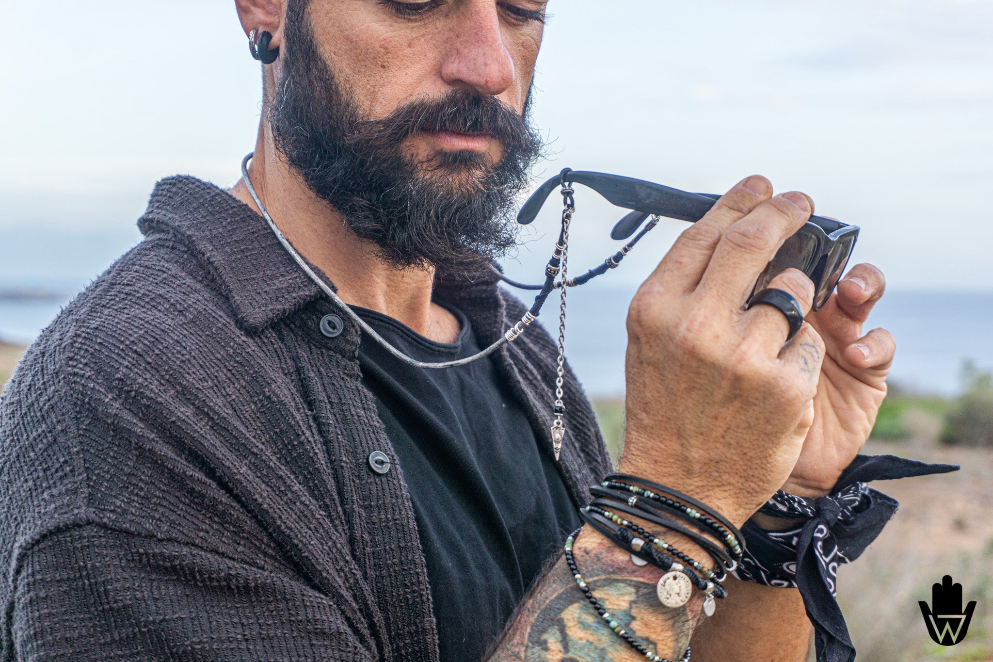A close-up lifestyle shot of a bearded man outdoors holding a black folding knife in his hands, wearing a dark textured button-up shirt, layered necklaces, and bracelets.