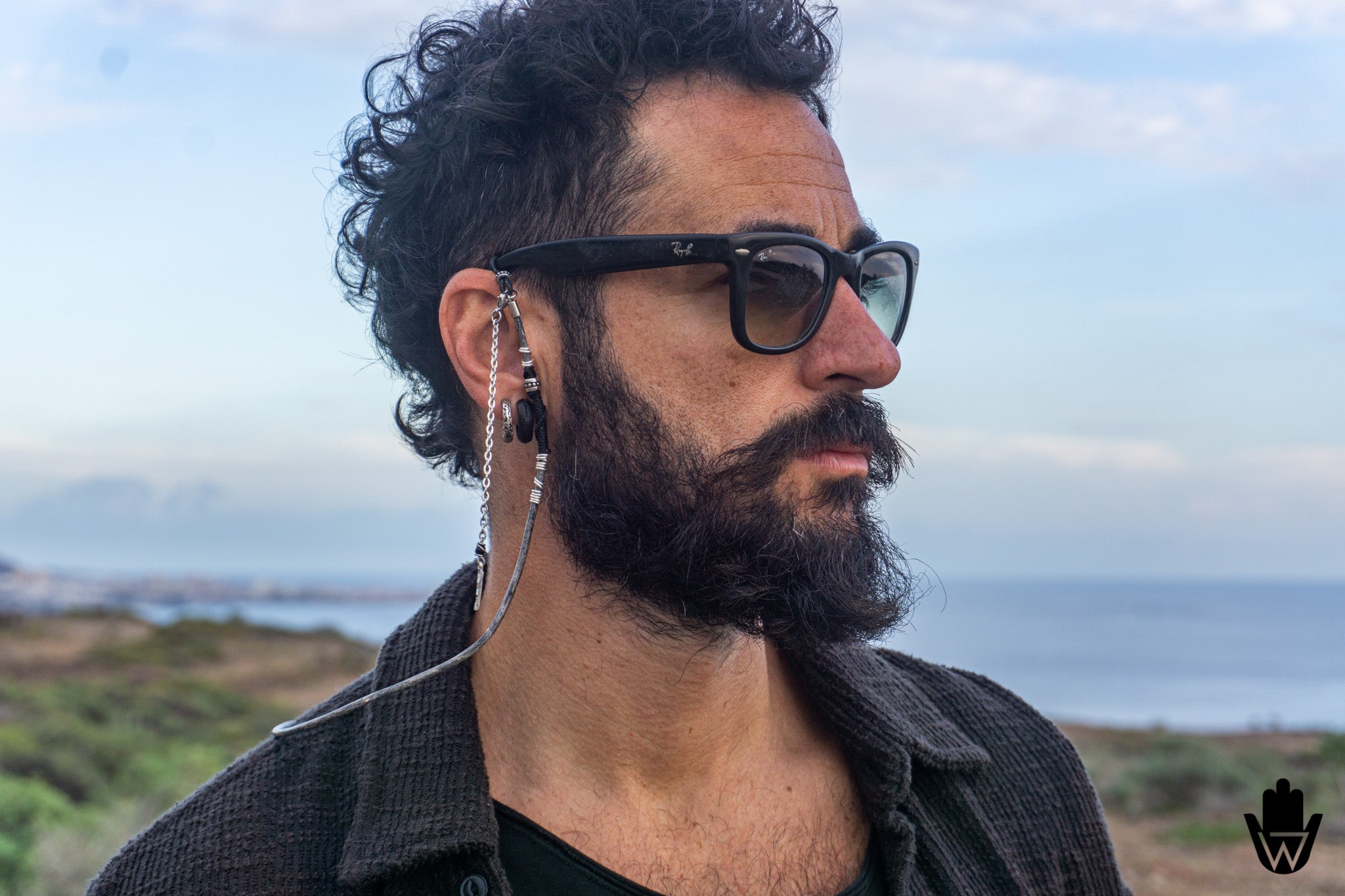 A man with a dark curly beard and mustache wearing black square-framed glasses with a long silver chain earring, photographed outdoors against a blurred coastal background.