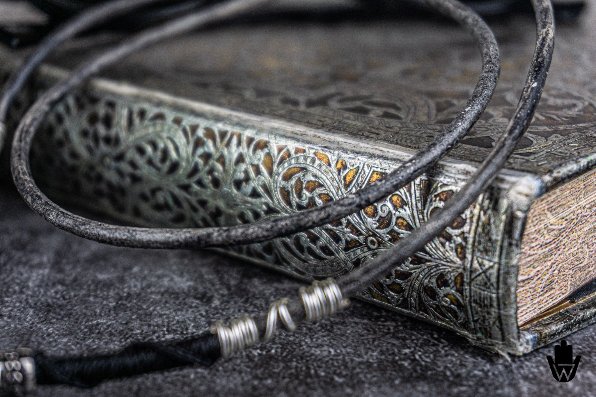 A close-up of a black cord necklace or bracelet with silver-toned wire-wrapped beads, draped over an ornate antique-style book on a dark textured surface.