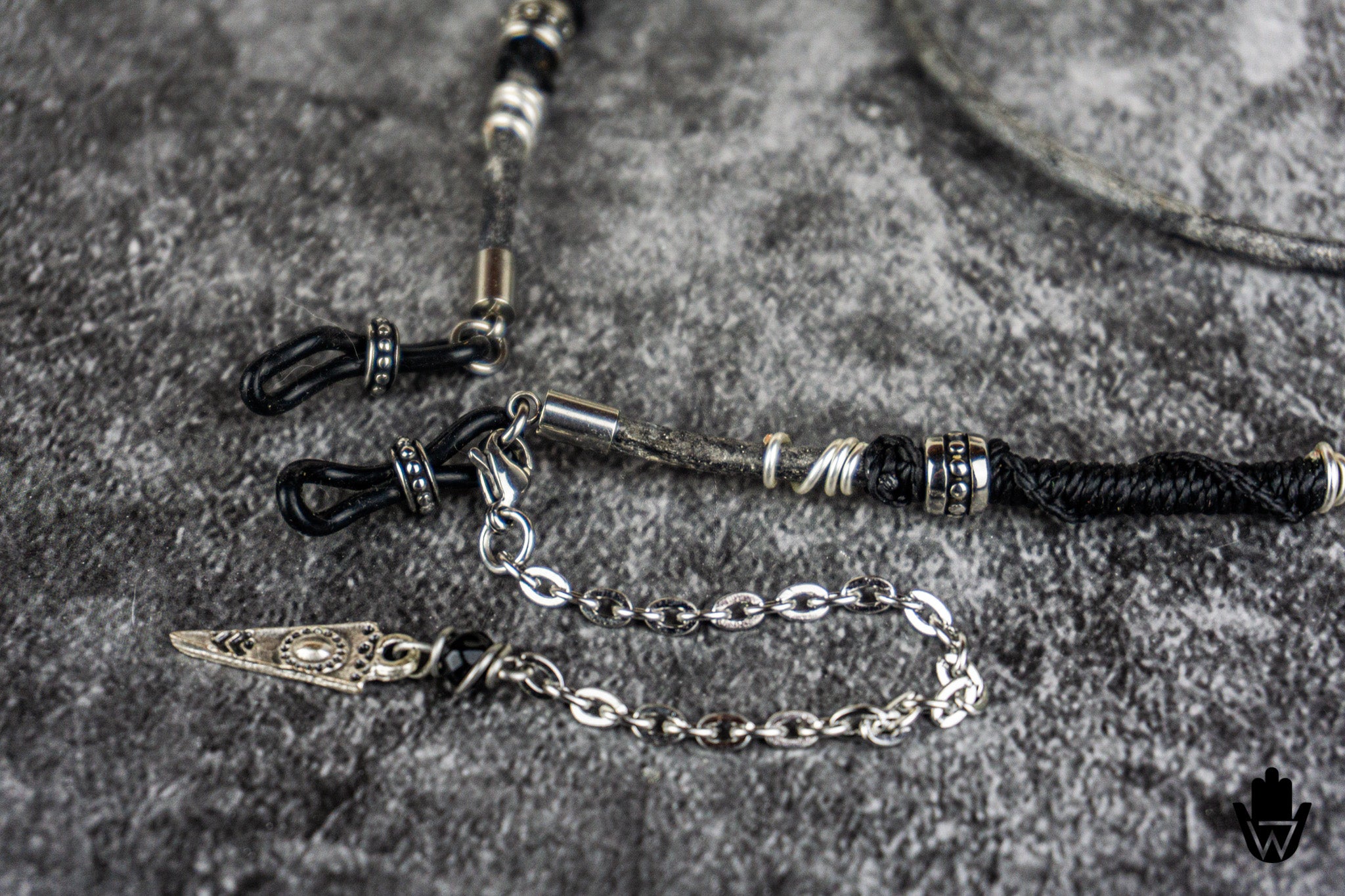 A close-up of a black braided cord necklace or bracelet with silver-toned chain links, decorative metal beads, and a small silver-toned pendant, laid on a textured gray surface.