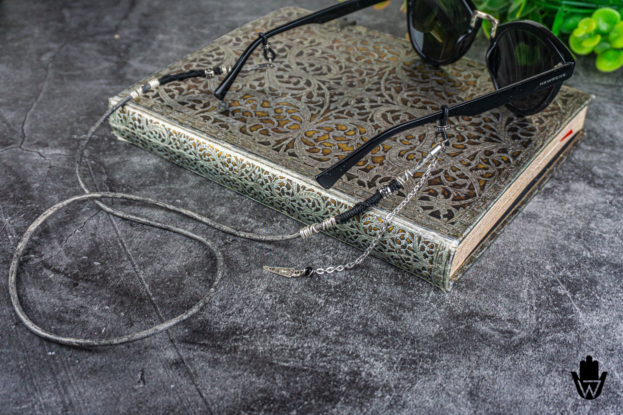 Black cat-eye sunglasses with silver chain and cord accessories resting on an ornate silver book over a dark gray textured surface.