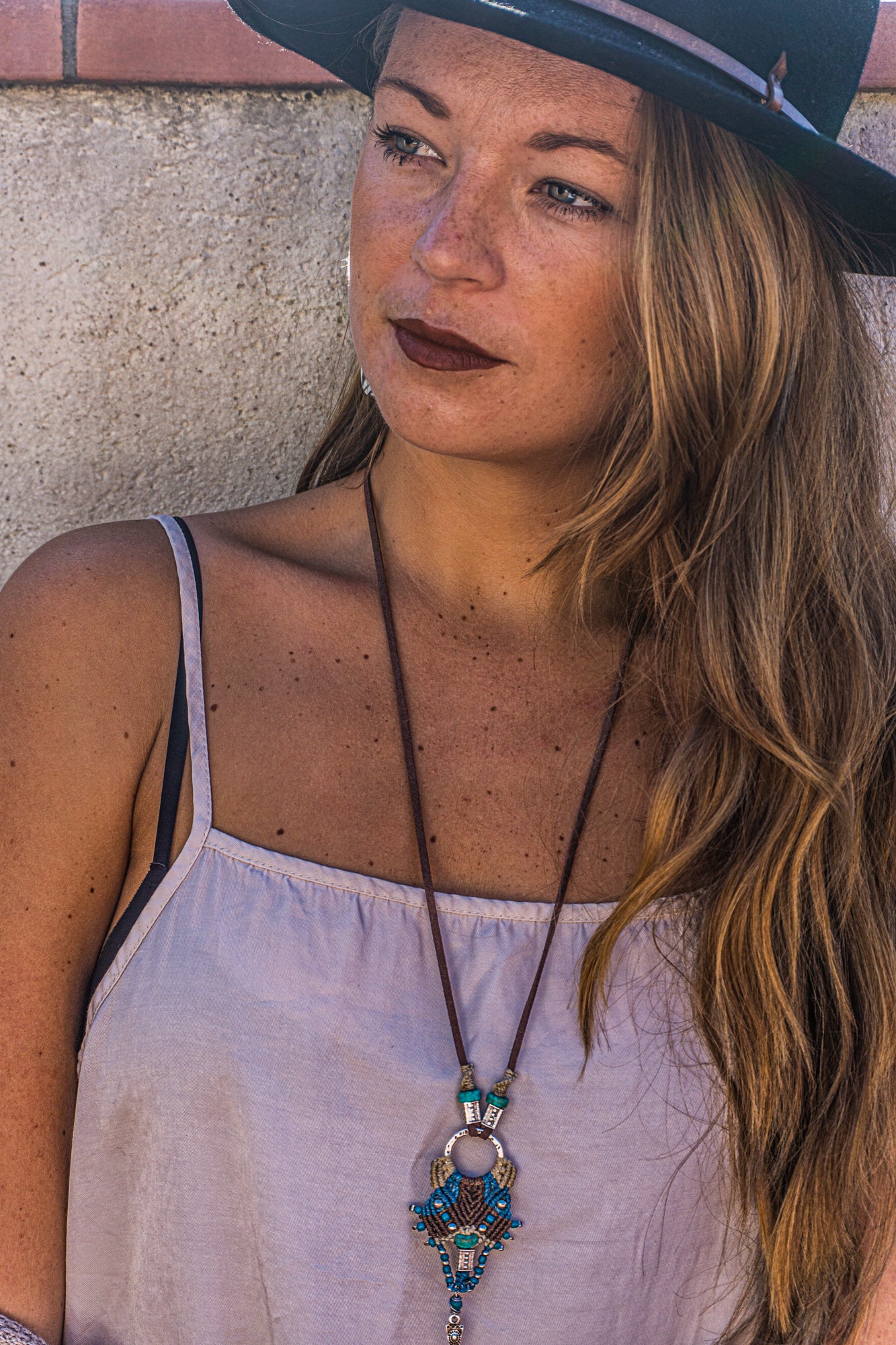 Woman wearing a hat and a necklace with a pendant against a stone wall.