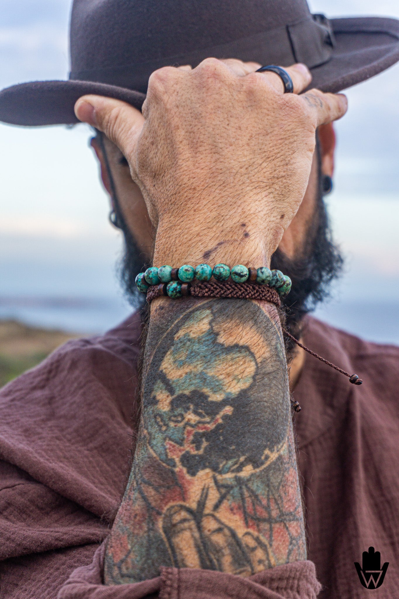 Person wearing a hat and brown jacket with a tattooed arm, holding a hat, with a macrame beaded bracelet set , against a blurred natural background.
