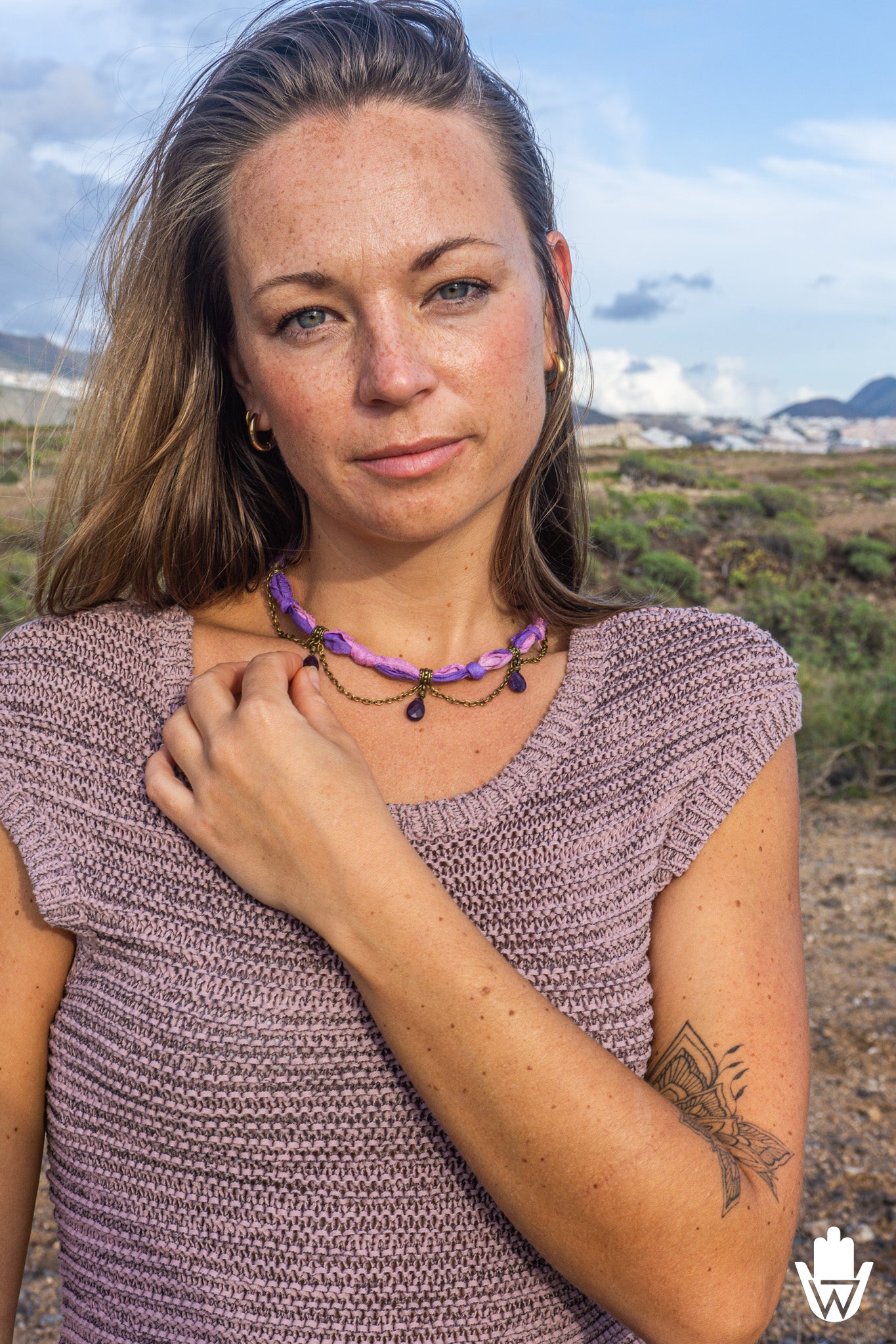 A woman wearing a textured mauve knit top and a purple beaded necklace with dark beads, posing outdoors against a blurred landscape background.
