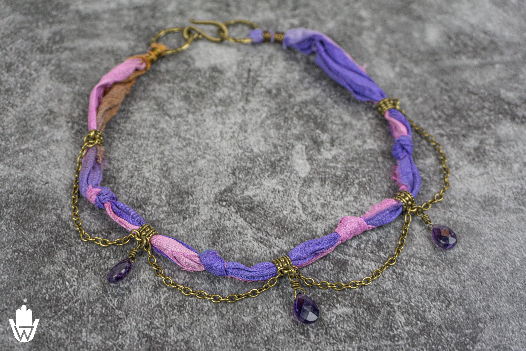 A handmade bohemian-style necklace featuring knotted purple and pink fabric strips, gold-toned chain links, and dangling amethyst teardrop charms, displayed on a textured gray background.