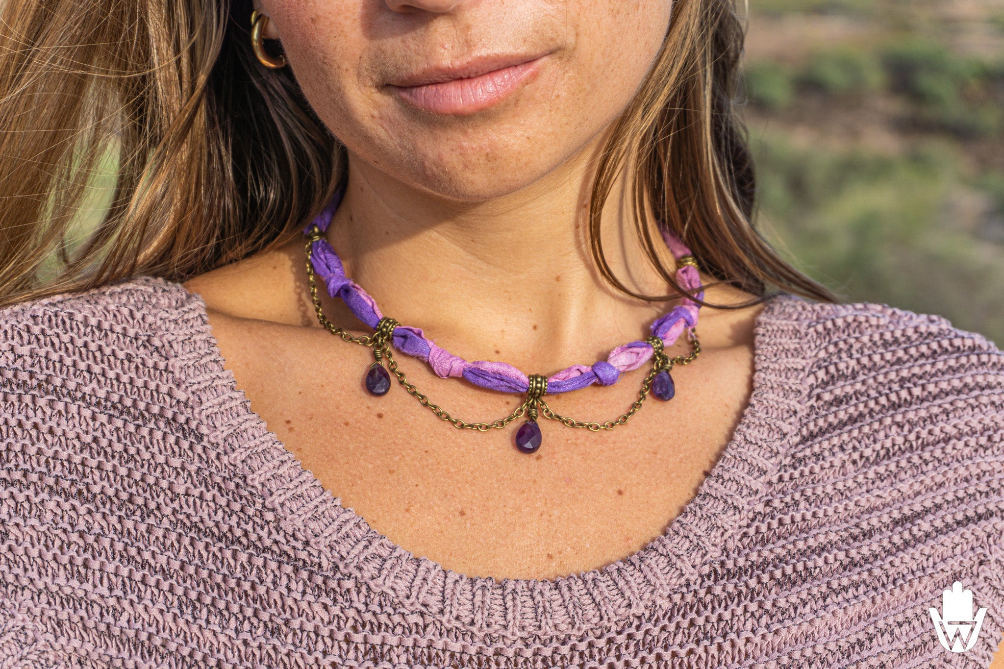 A close-up of a woman wearing a bohemian-style purple and bronze chain necklace with teardrop charms, paired with a mauve knit sweater.