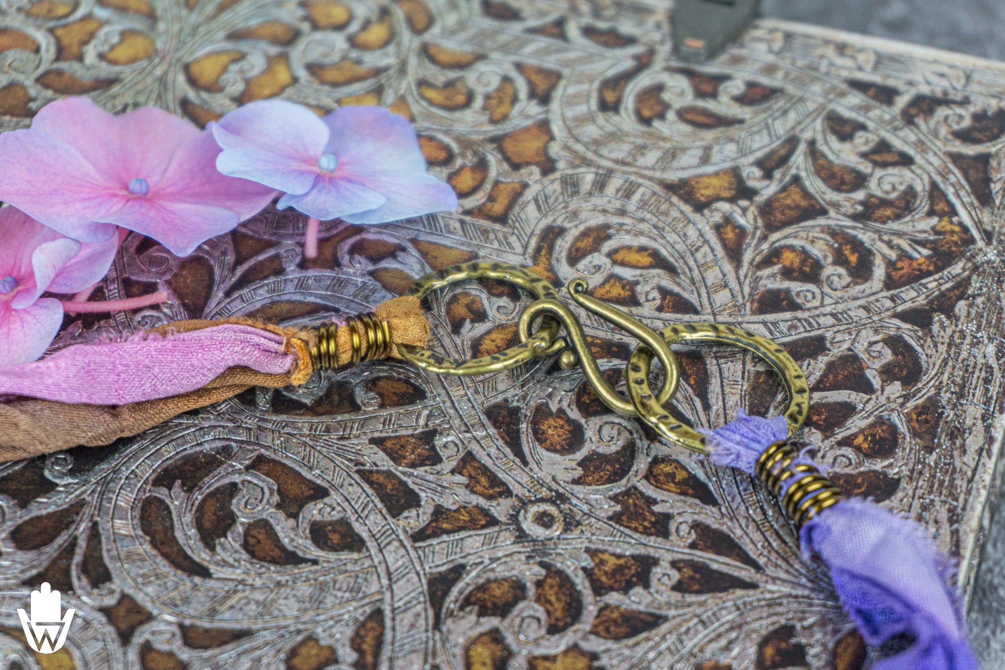 A close-up of a decorative jewelry piece featuring a gold-toned interlocking ring clasp, wrapped with pink and purple fabric, and accented with gold wire beads, resting on an ornate brown patterned surface with pink flowers.