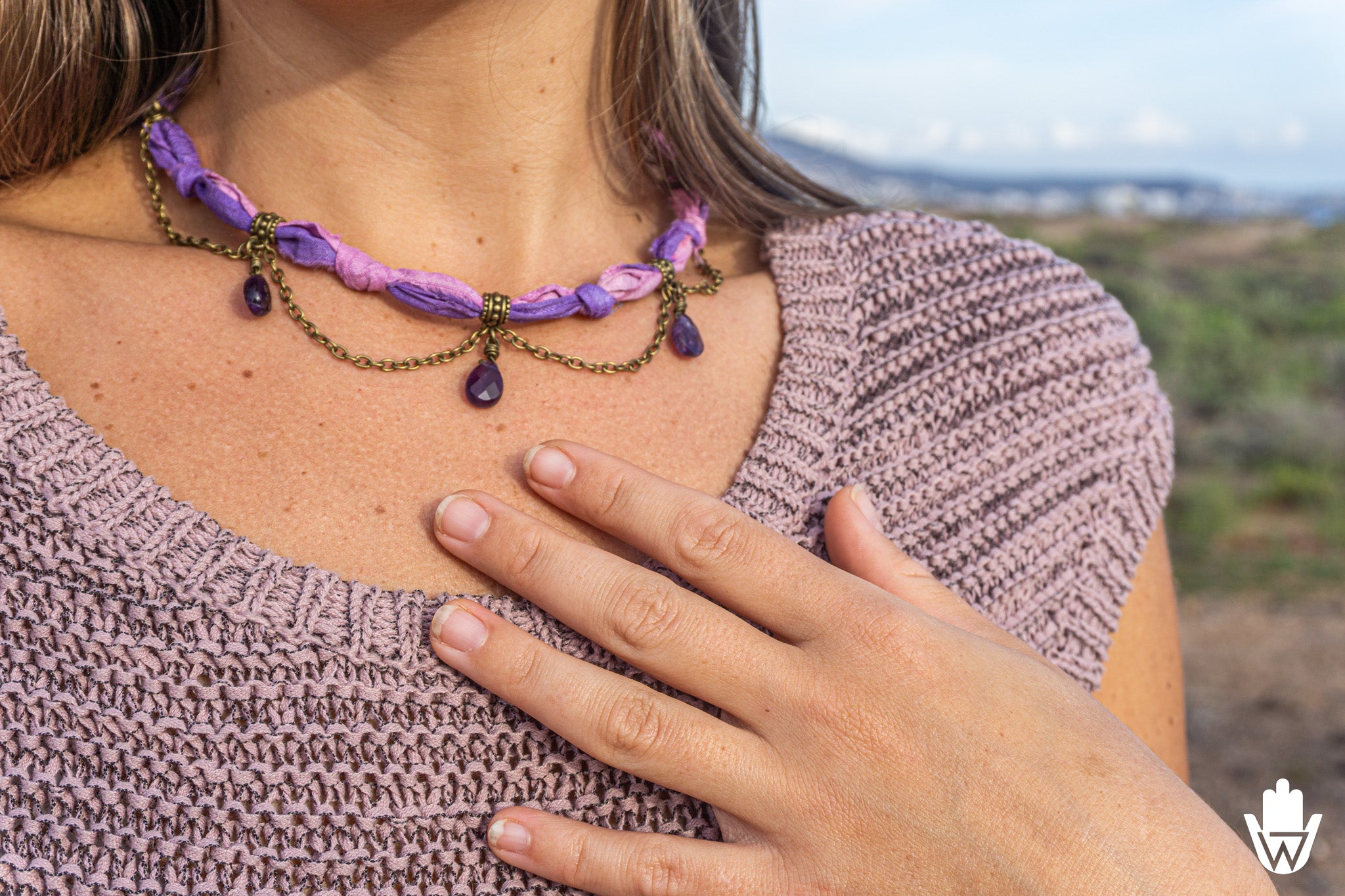 A close-up lifestyle shot of a woman wearing a bohemian-style necklace with purple fabric loops, bronze chain links, and purple teardrop gemstones, set against an outdoor blurred background.