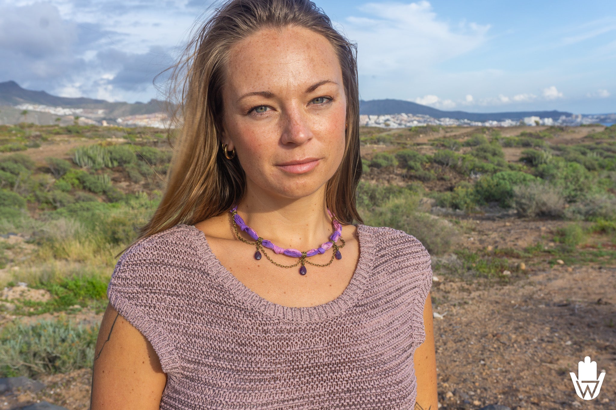 A woman with blonde hair wearing a purple beaded necklace and a mauve knit top, standing outdoors in a dry, scrub-covered landscape with mountains and a cloudy sky in the background.