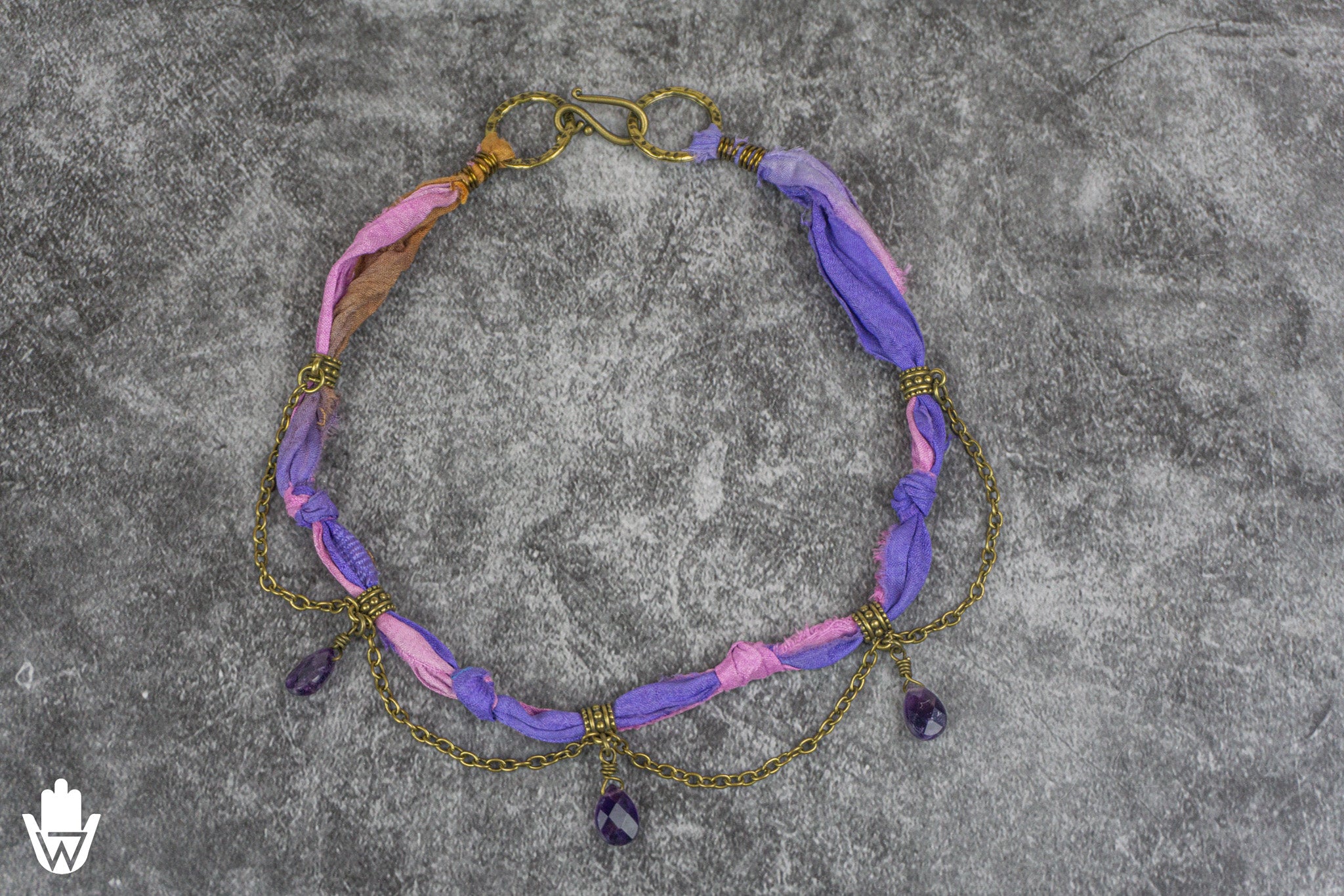 A handmade statement necklace featuring knotted purple and pink fabric strands, draped with gold-toned chains and dangling amethyst beads, displayed on a textured gray background.