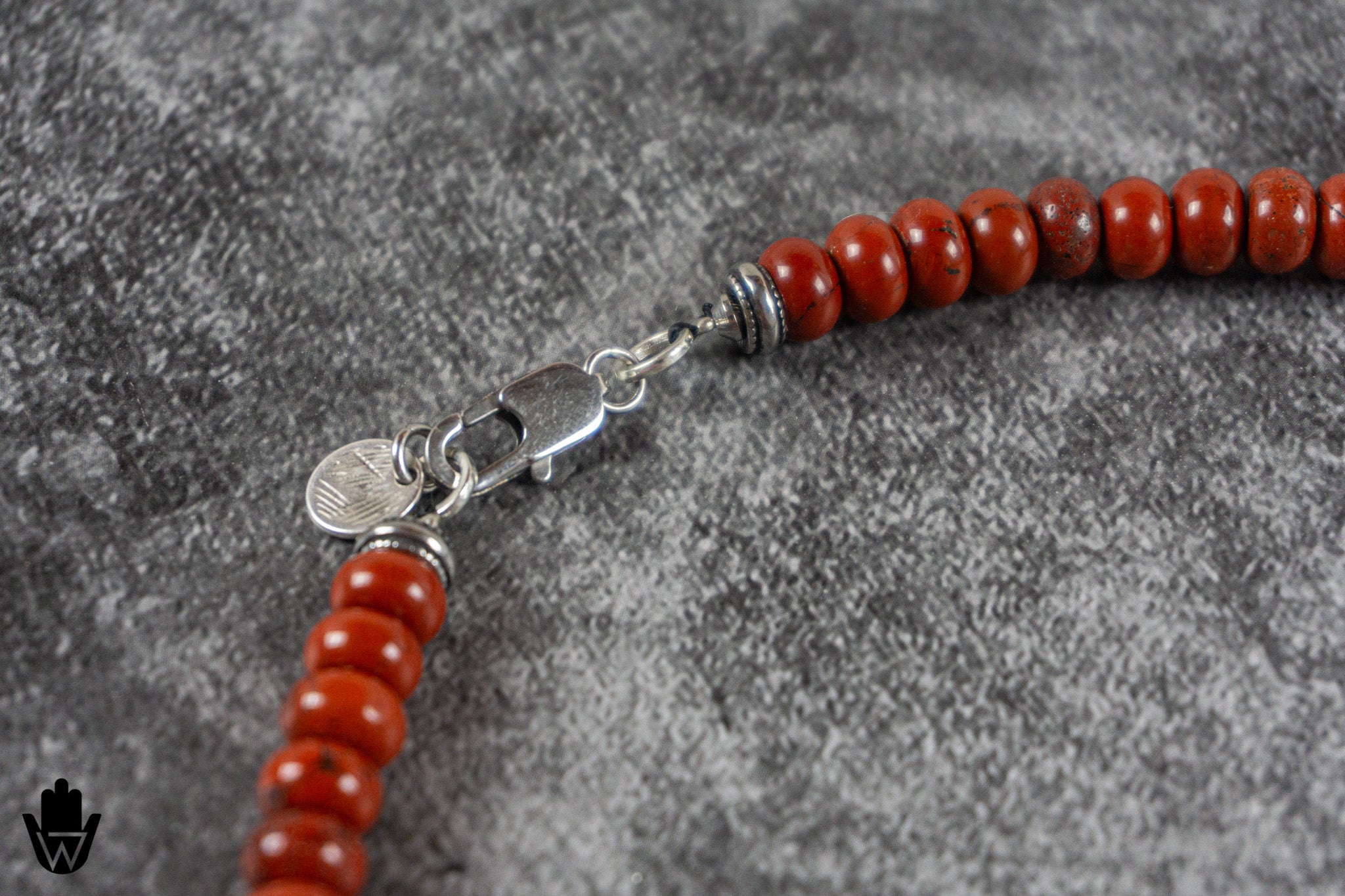 A close-up of a beaded necklace featuring round, polished reddish-orange beads and a silver-toned lobster clasp, displayed on a textured gray background.