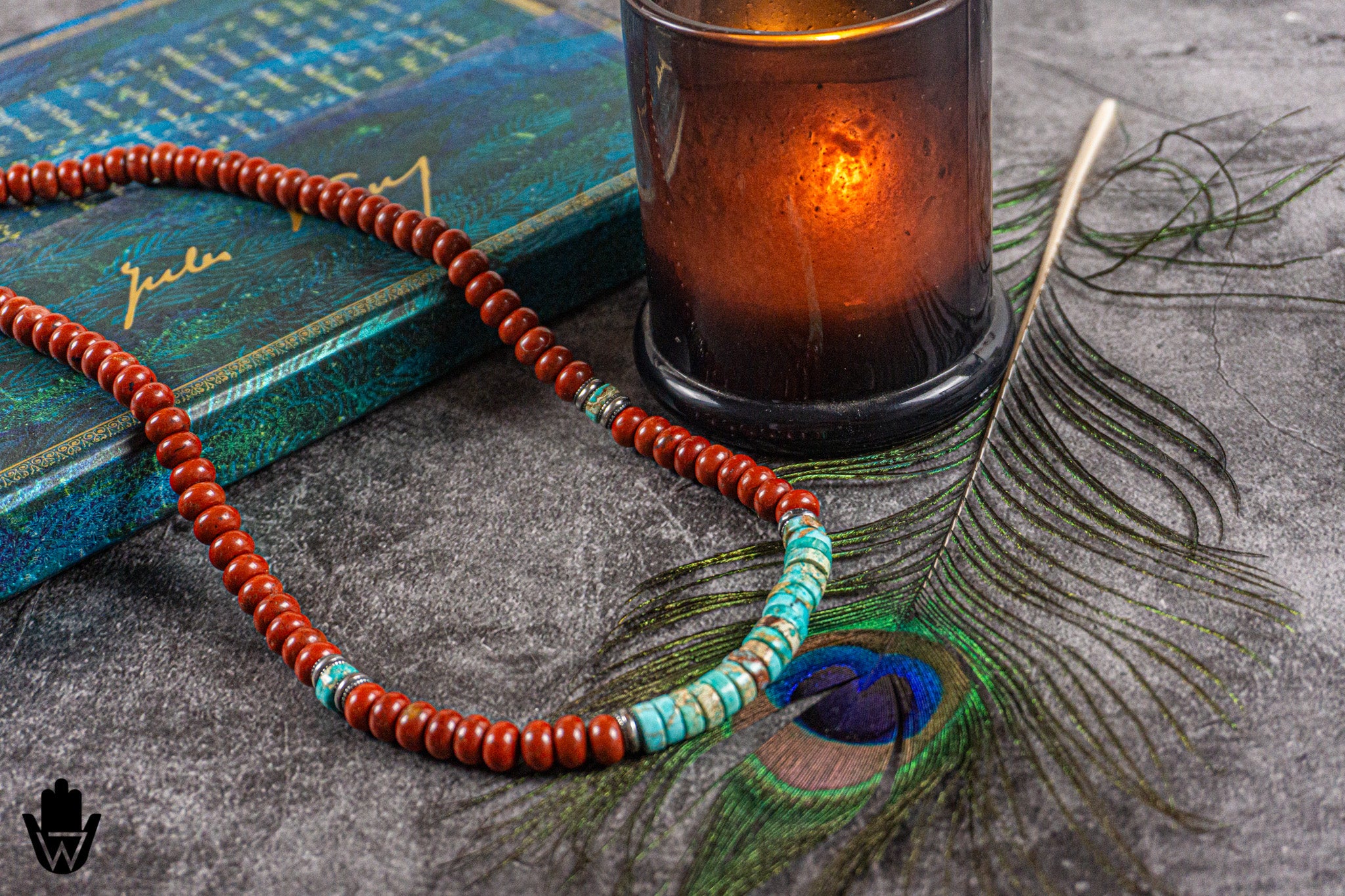 A beaded necklace with red-orange round beads and turquoise accents, styled on a textured gray surface next to a teal book, a glowing amber candle, and a peacock feather.