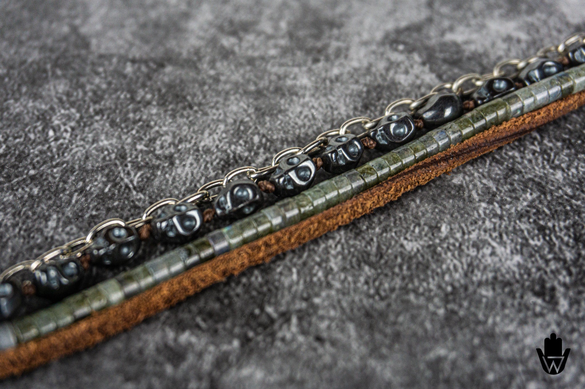 A close-up of a beaded leather strap or bracelet featuring dark metallic chain links, polished black beads, textured green-gray beads, and a brown leather base, laid diagonally on a gray textured surface.
