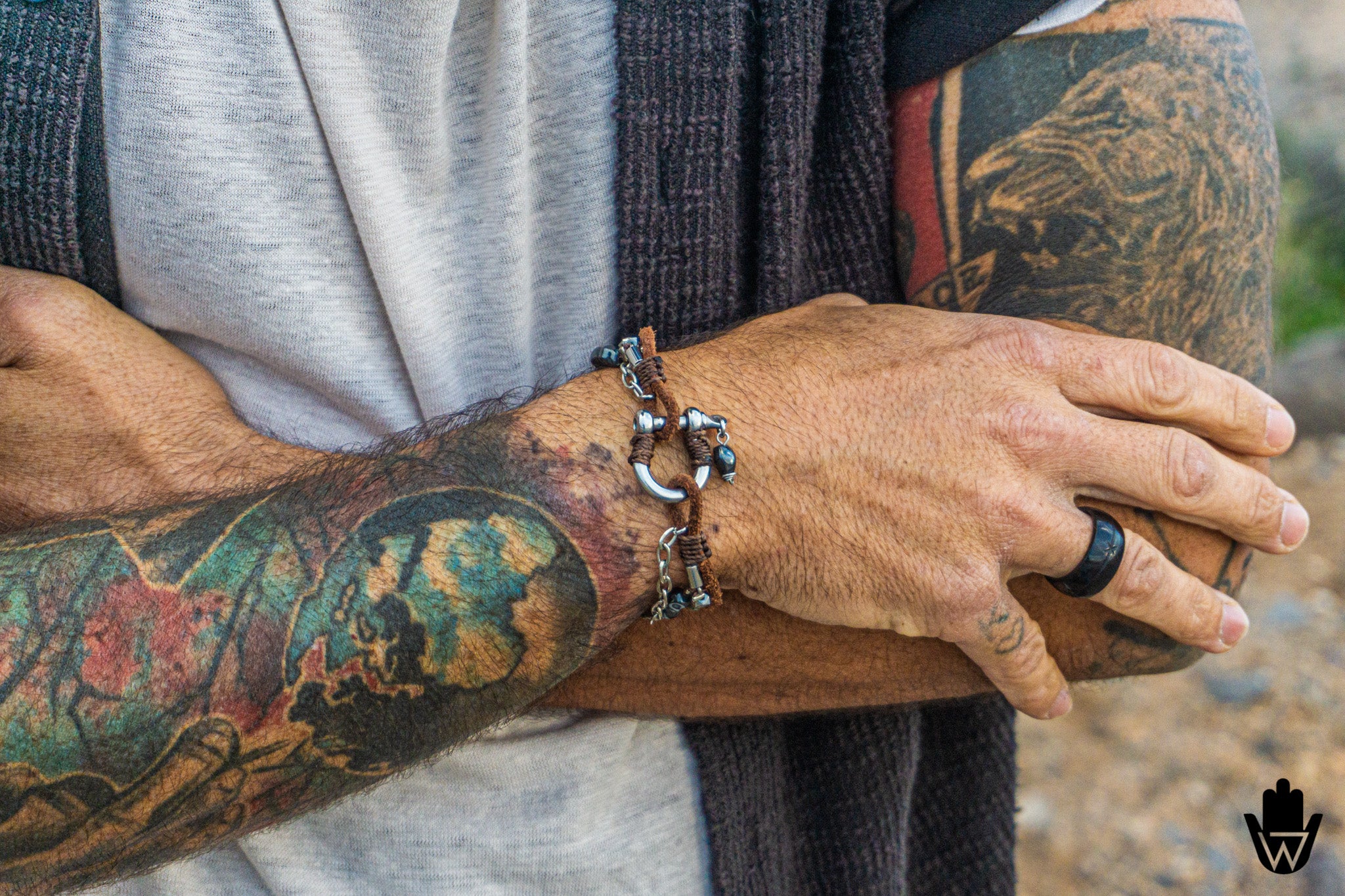 A close-up of a person's tattooed arms crossed, showcasing a dark metal chain bracelet with silver-toned charms on the wrist. The person is wearing a gray t-shirt, a dark gray knit cardigan, and a black ring.