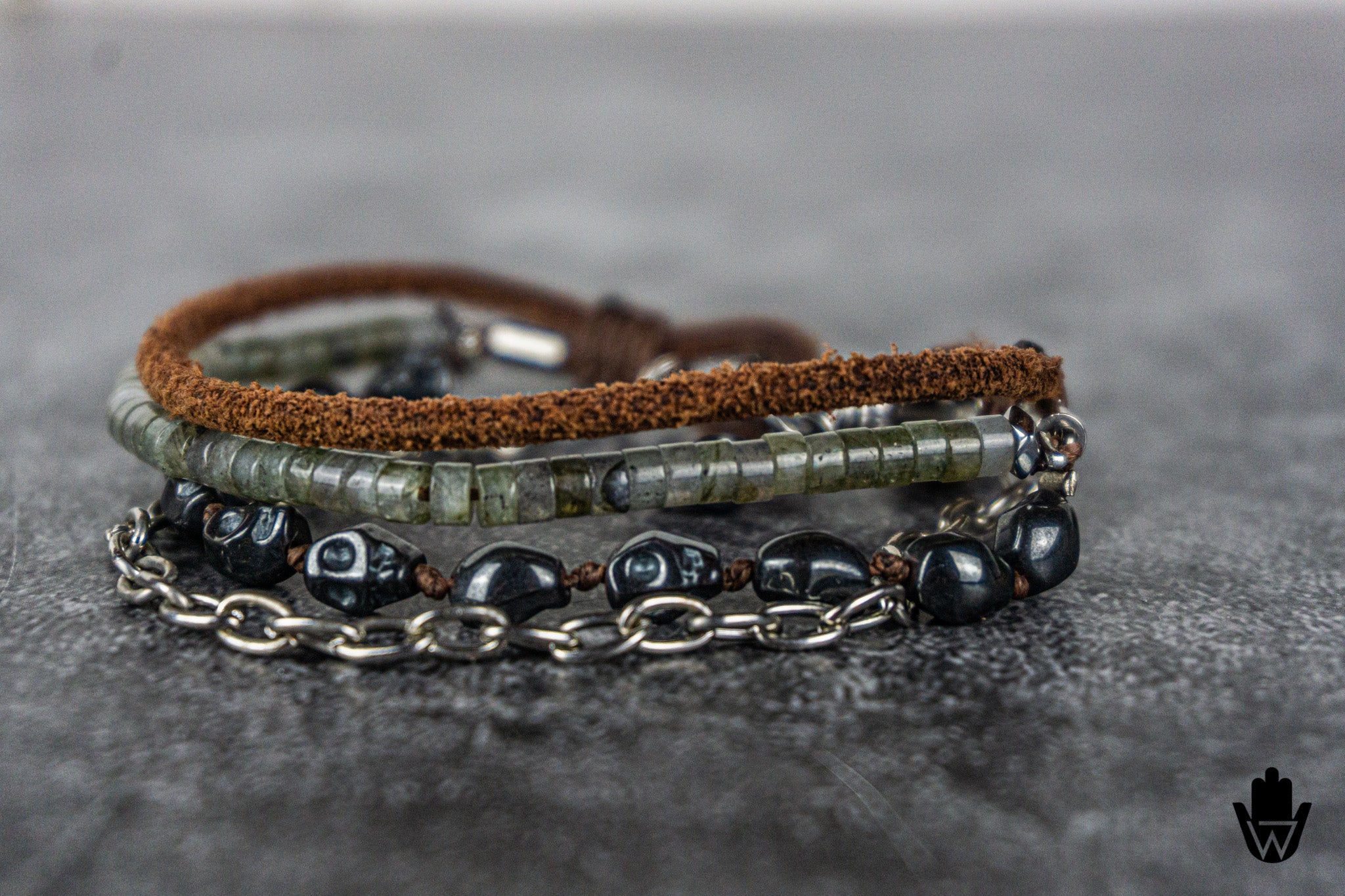 A close-up of a multi-layered bracelet featuring a brown suede-like band, a strand of green-gray labradorite beads, and a silver chain with dark skull beads, resting on a textured gray surface.
