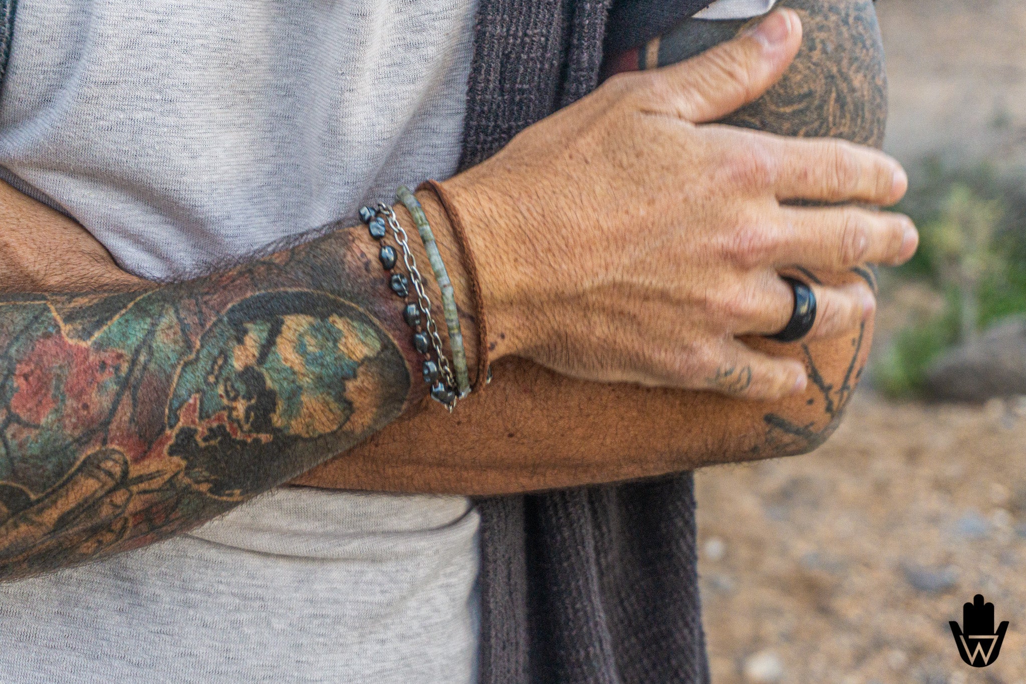 Close-up of a tattooed person's crossed arms, showcasing a stack of bracelets on the left wrist and a black ring on the right hand, with a gray t-shirt and dark knit sweater visible.