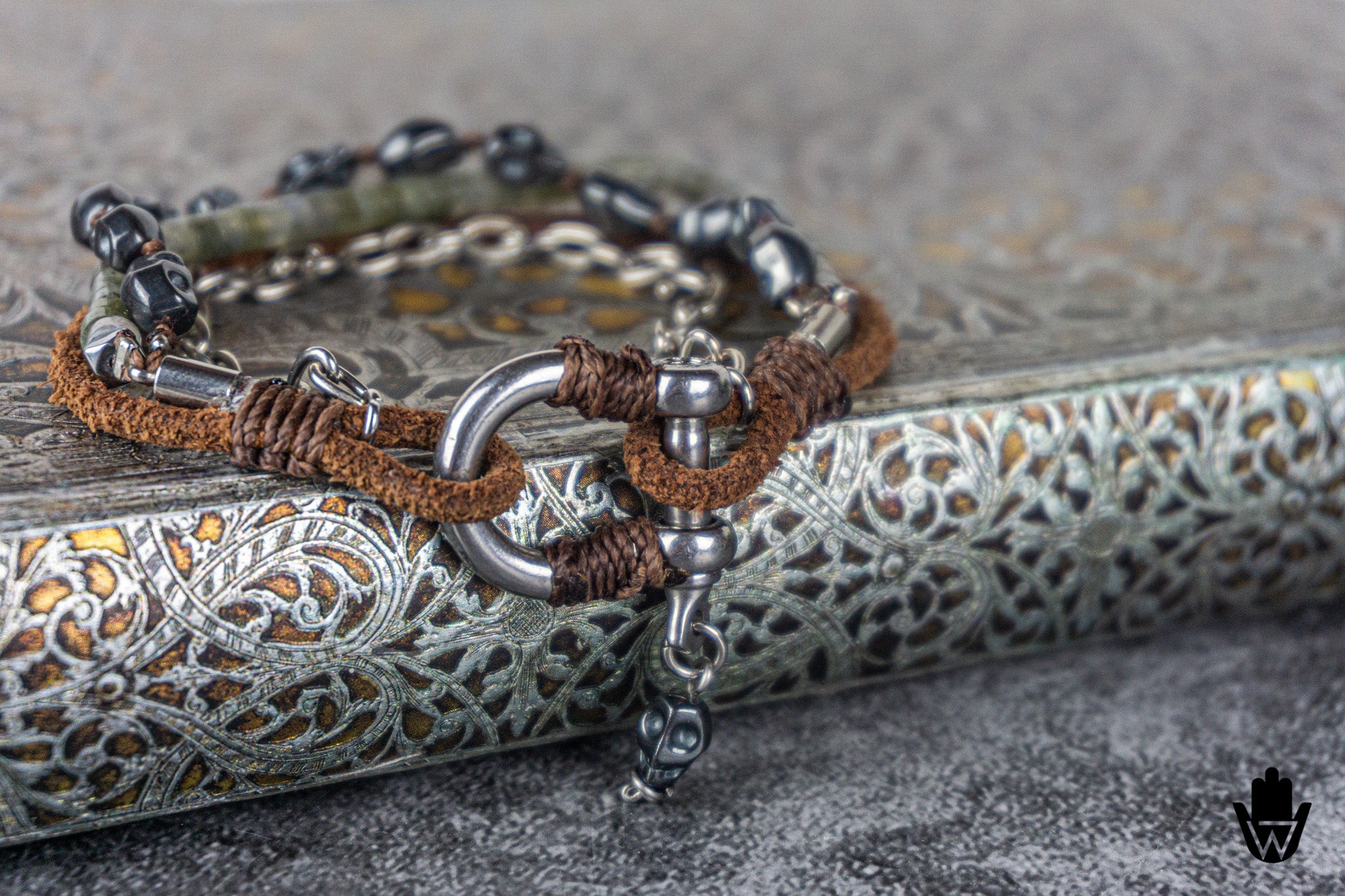A close-up of a multi-strand bracelet featuring brown suede, dark metallic beads, silver-toned metal rings, and a small hanging silver charm, displayed on an ornate silver book.