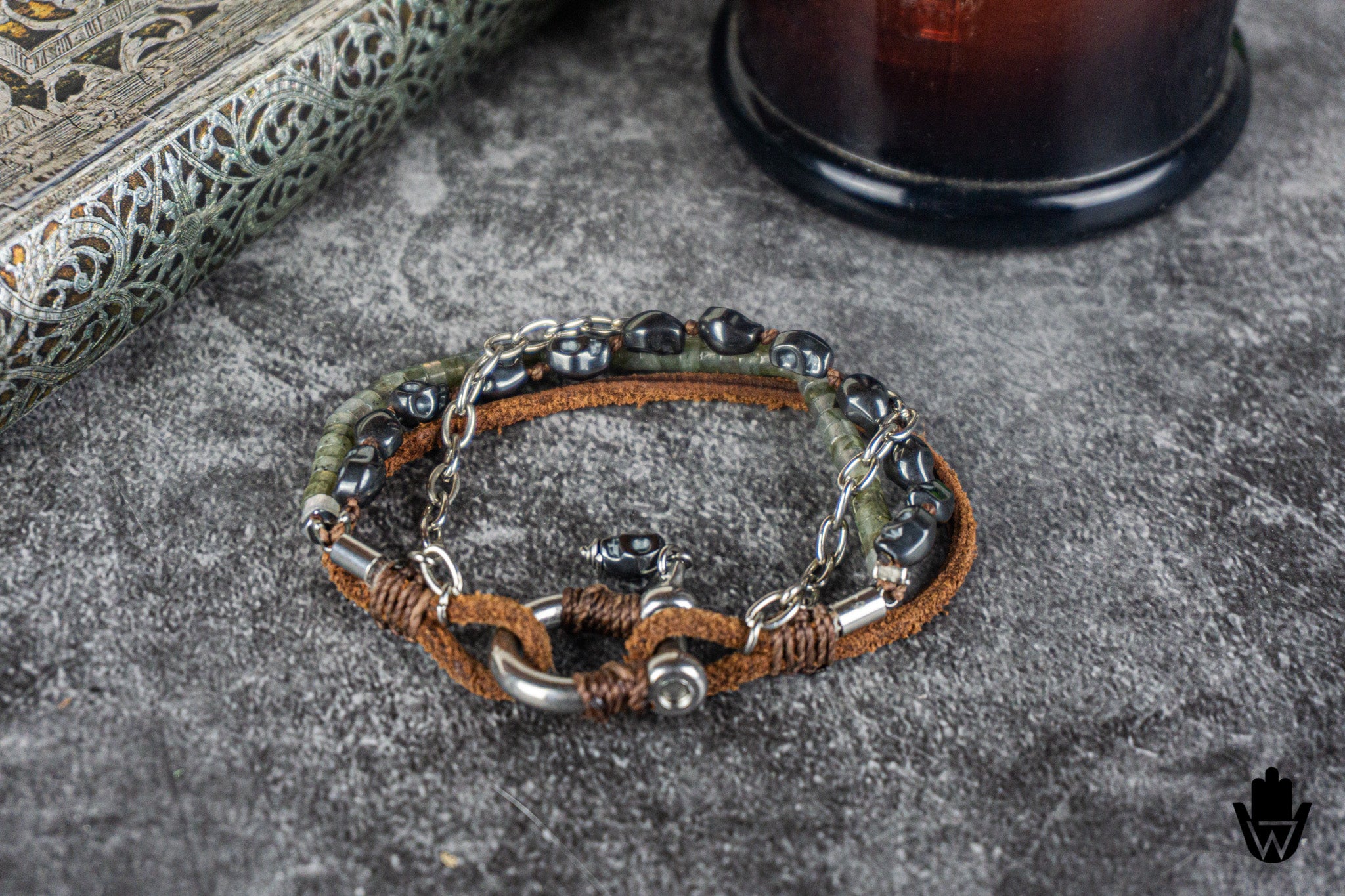 A double-layered men's bracelet featuring brown leather cords, dark metallic beads, silver-toned chain links, and a central silver-toned chain with a small black bead accent, displayed on a textured gray stone surface.