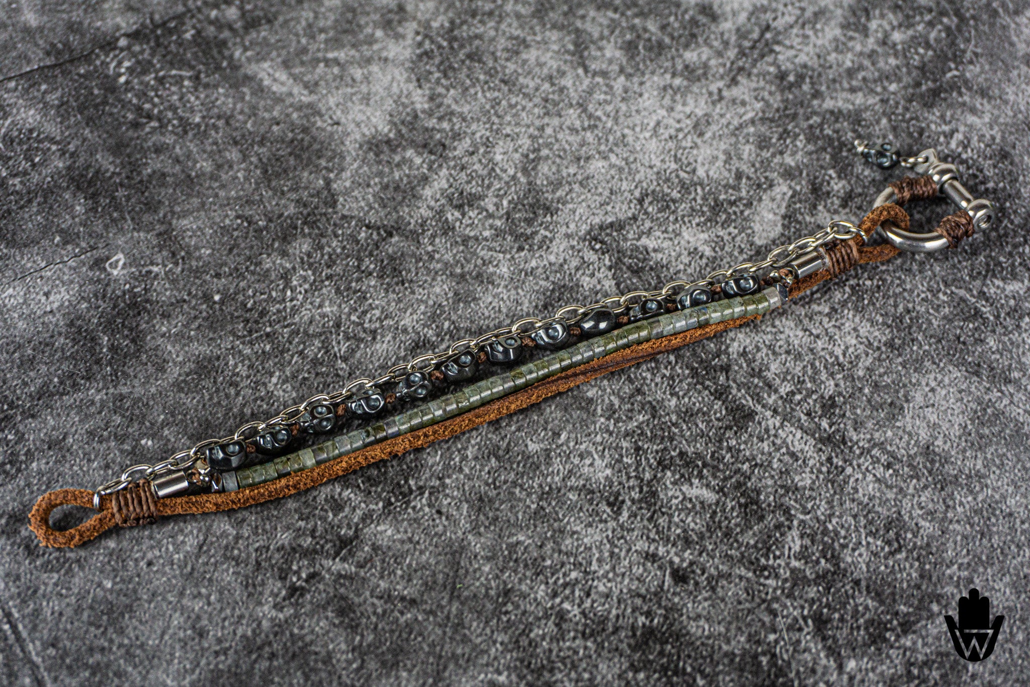 A handmade bracelet featuring a brown leather cord, a central silver-toned chain with dars kull beads, and a green stone inlay, laid diagonally on a textured gray background.