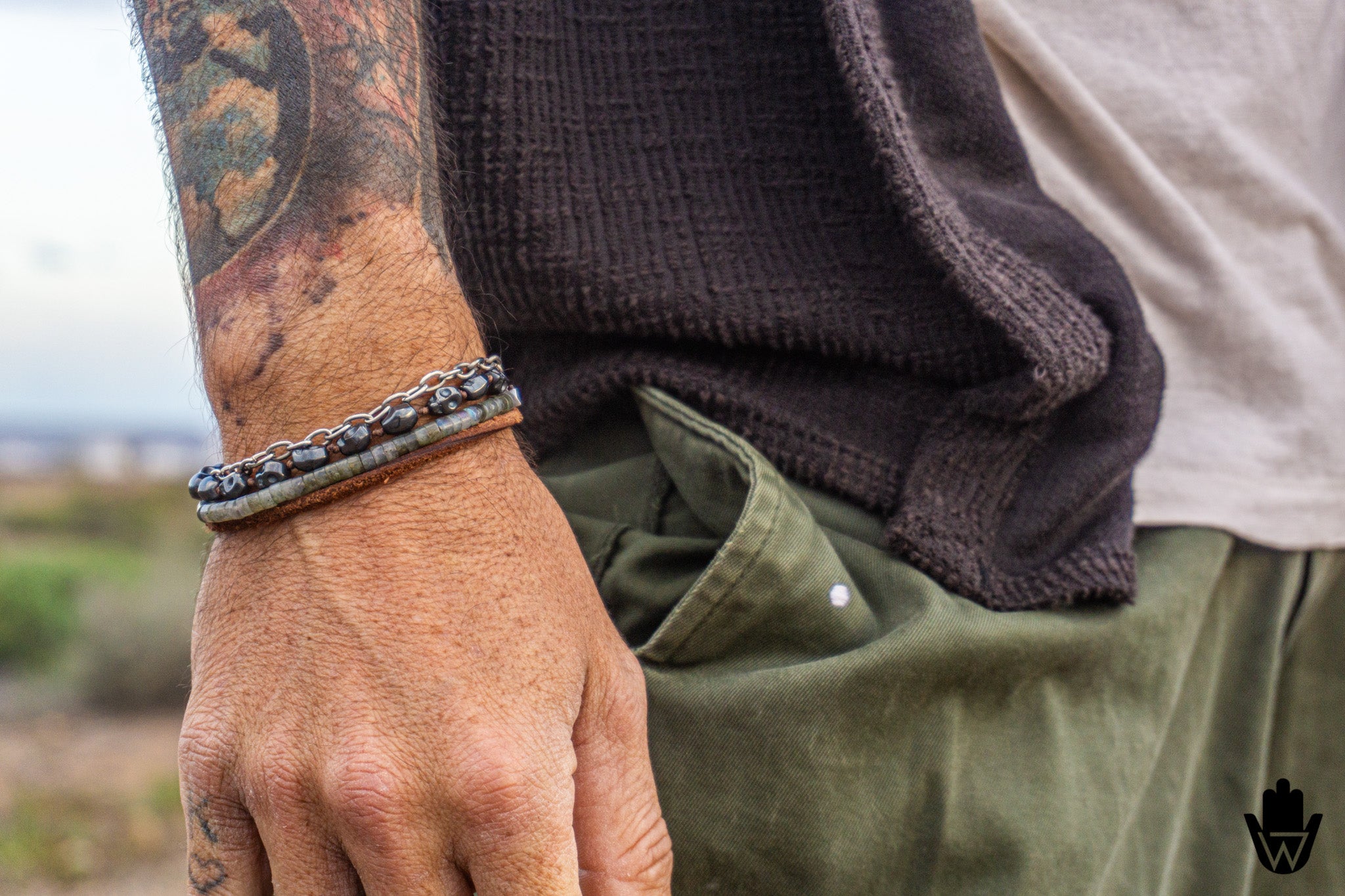 A close-up of a person's tattooed forearm and hand wearing a beaded bracelet with dark metallic chain links, styled with a dark knit sweater and olive green pants.