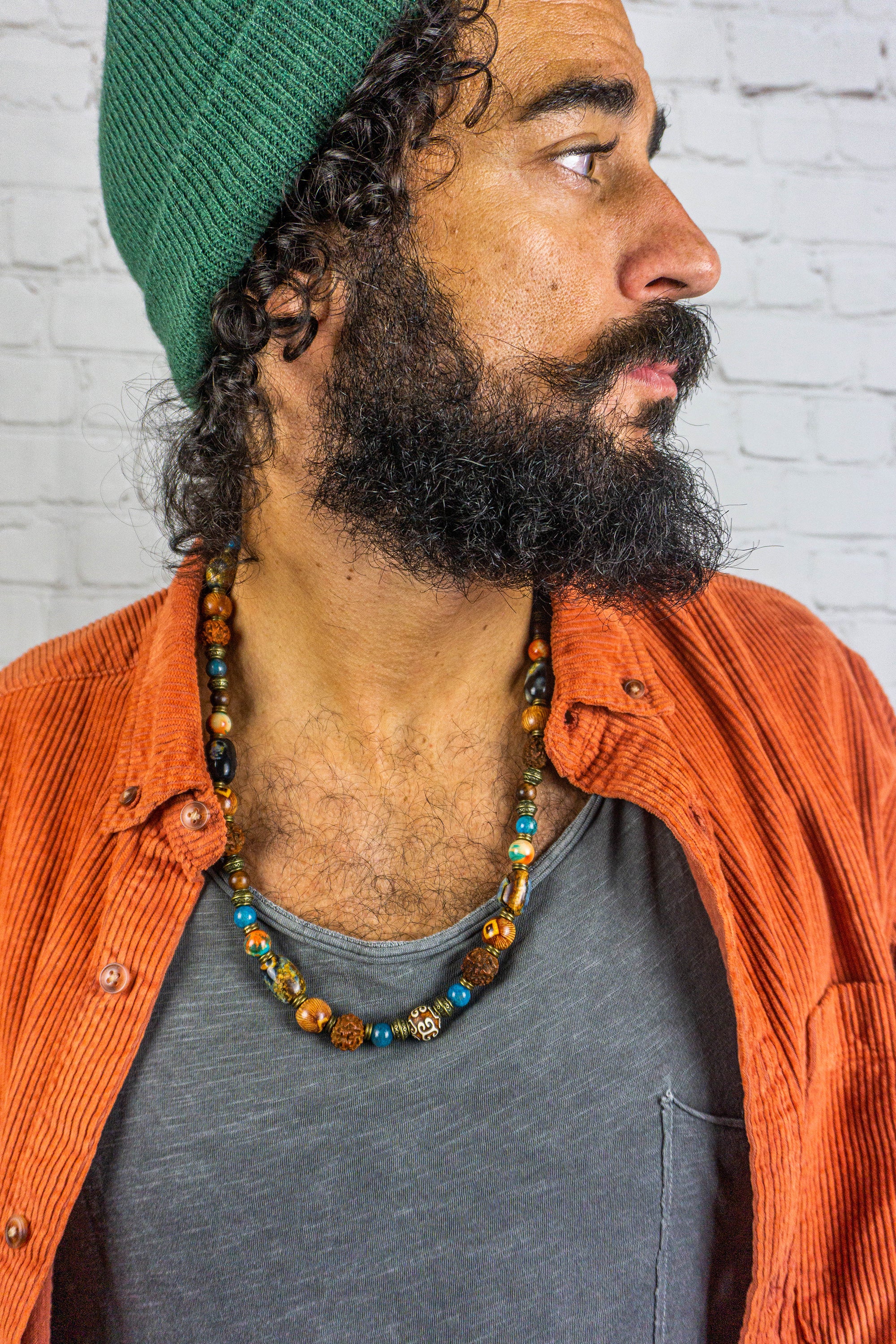 Man wearing a green beanie, orange jacket, and gray shirt with a colorful necklace against a white brick wall.