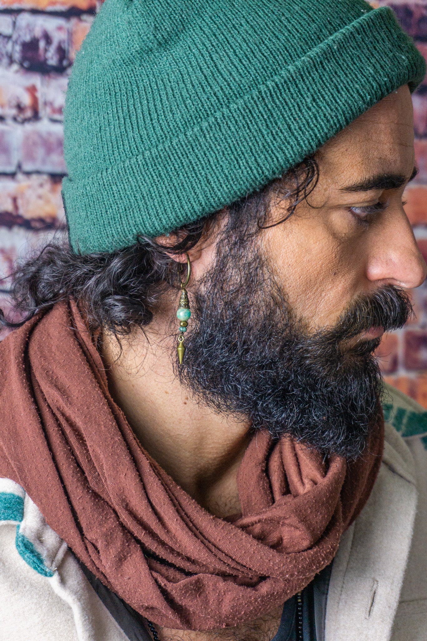 A man with a green beanie, brown scarf and a dangling earring with a green stone and a little bronze spike- wander jewellery